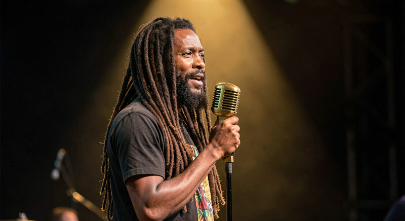 A reggae singer with dreadlocks at a vintage microphone in red gold and green stage haze.