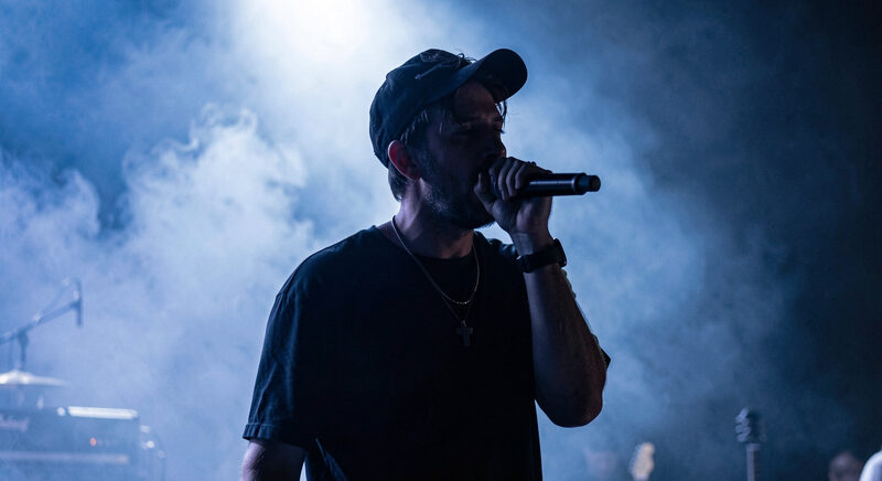 A male singer with a cap performing on a dark trip hop stage crossed by blue and red light beams.
